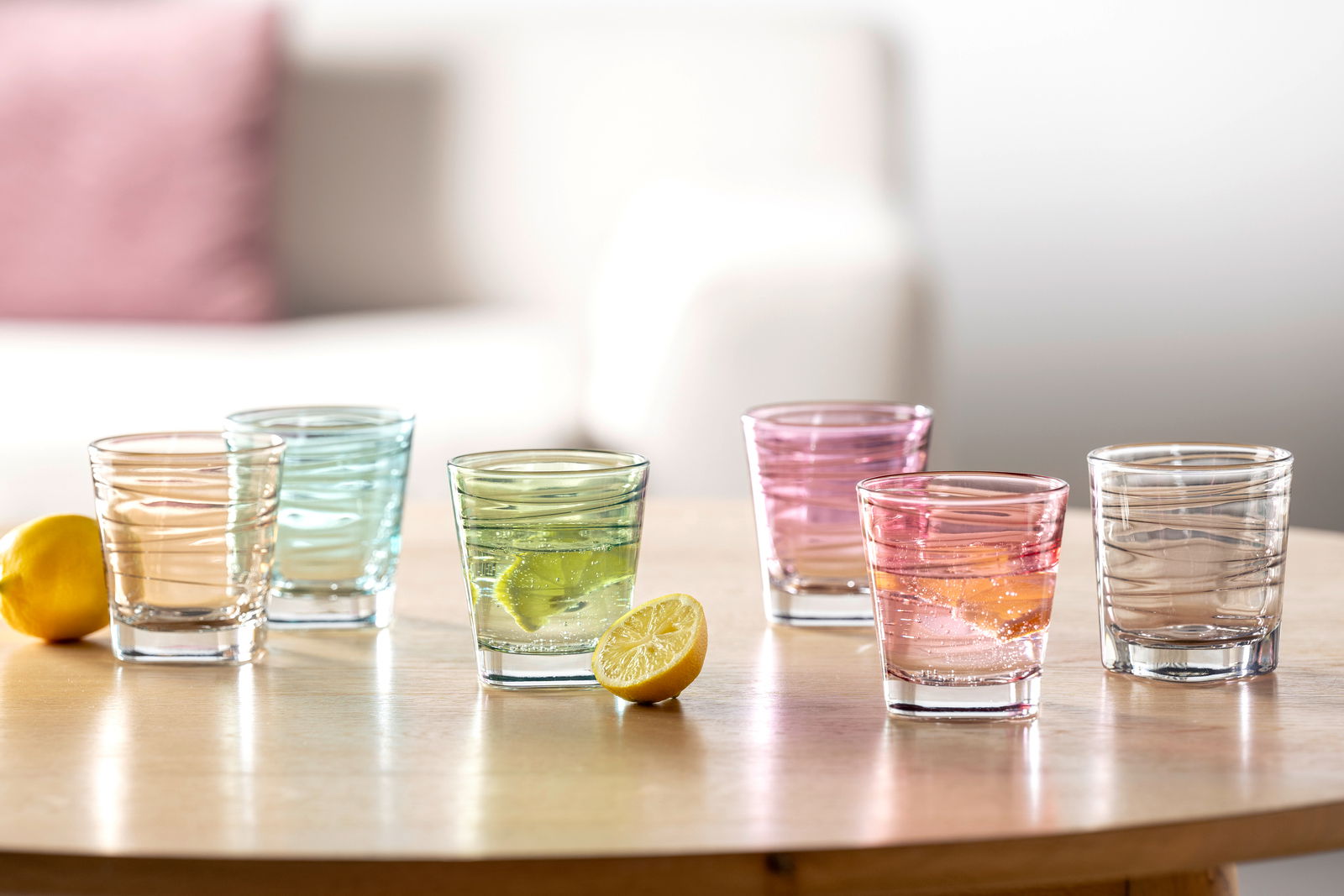 Sechsteiliges Becherset mit 250 ml Fassungsvermögen in verschiedenen Farben, auf einem Tisch arrangiert, mit einer Zitrone im Vordergrund. Perspektive: seitlich.