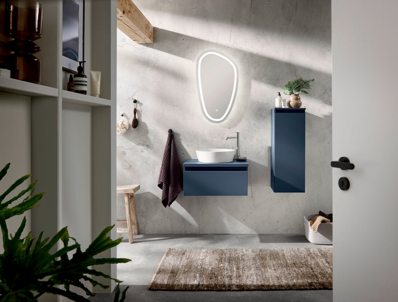 Moderne Badkombination mit blauem Waschtisch und Hängeschrank, beleuchtetem Spiegel und Waschbecken aus der Perspektive einer offenen Tür.