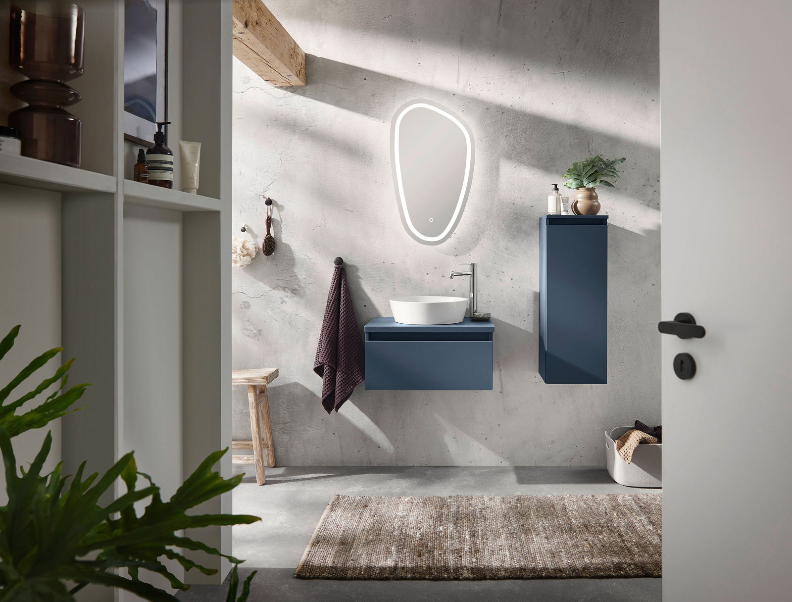 Moderne Badkombination mit blauem Waschtisch und Hängeschrank, beleuchtetem Spiegel und Waschbecken aus der Perspektive einer offenen Tür.