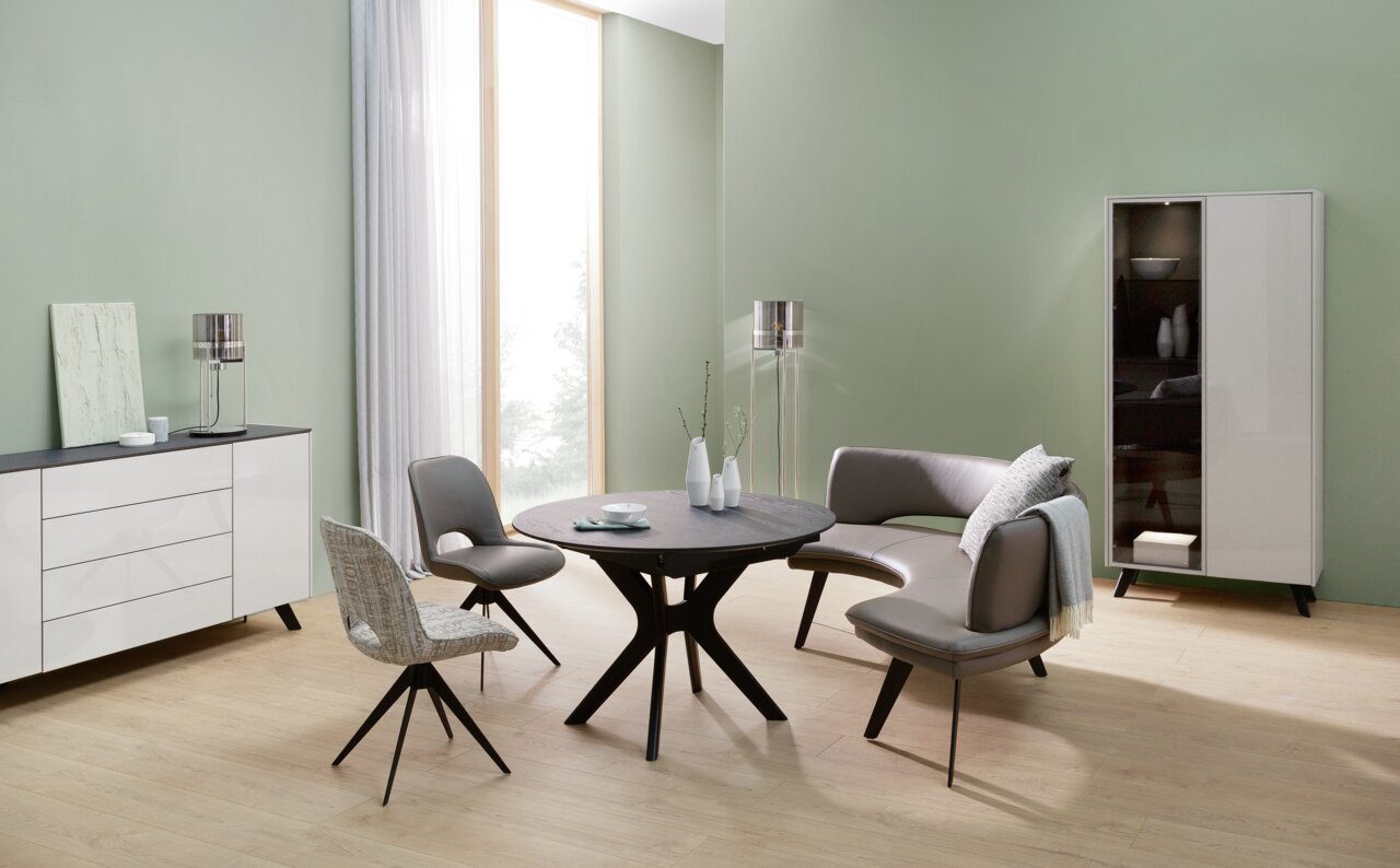 Moderner Esstisch in einem stilvollen Wohnzimmer mit grüner Wand, umgeben von eleganten Stühlen und einem geschwungenen Sofa, aus der Frontalperspektive betrachtet.