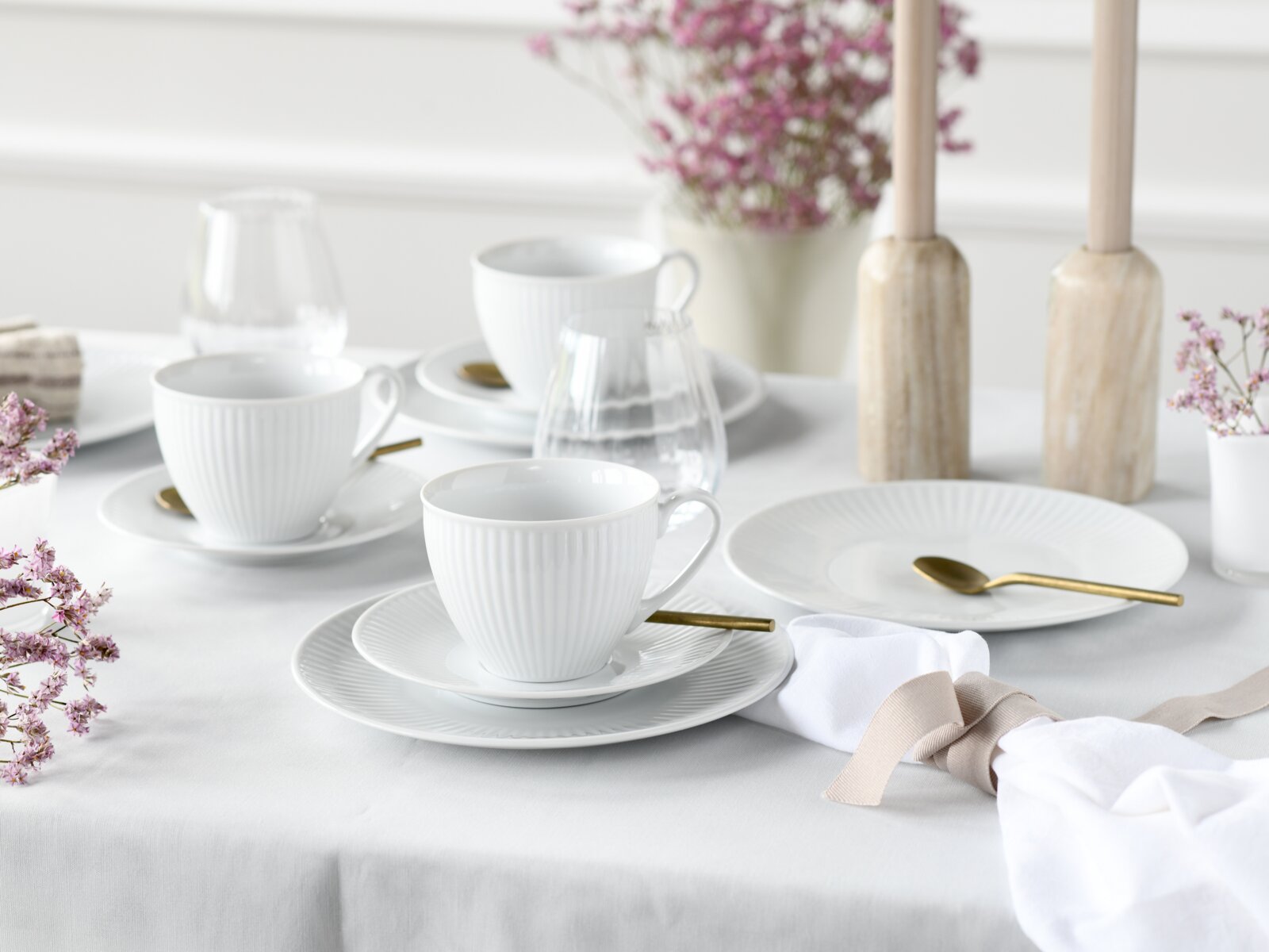 Schlichtes weißes Porzellan-Kaffeegeschirr mit gerippten Tassen, Untertassen und Tellern auf gedecktem Tisch mit goldfarbenen Löffeln, Gläsern und zarten Blumen, aufgenommen aus leicht erhöhter schräger Perspektive.