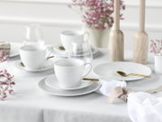 Schlichtes weißes Porzellan-Kaffeegeschirr mit gerippten Tassen, Untertassen und Tellern auf gedecktem Tisch mit goldfarbenen Löffeln, Gläsern und zarten Blumen, aufgenommen aus leicht erhöhter schräger Perspektive.