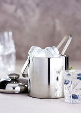Eiskübel aus Edelstahl mit Zange, gefüllt mit Eiswürfeln, aus seitlicher Perspektive fotografiert. Im Hintergrund ein Glas mit Getränk und Beeren.