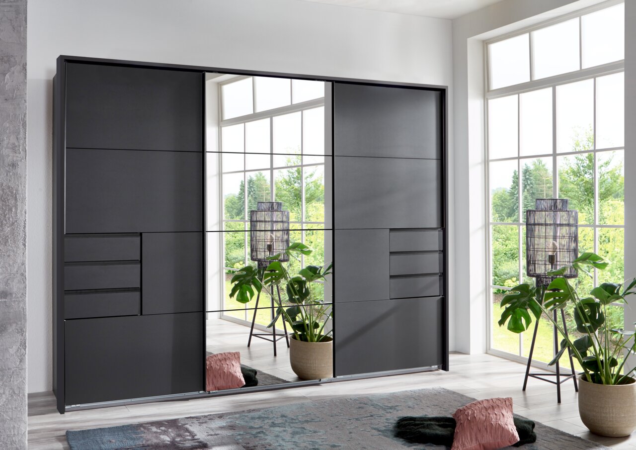 Z2 Schwebetürenschrank/Passp.3trg SAIGON Moderner Schwebetürenschrank in dunklem Design mit Spiegel in einem hellen Raum, seitliche Perspektive.