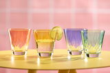 Vier bunte Trinkgläser auf einem gelben Tisch vor einem rosa Hintergrund, aus der Vorderansicht fotografiert.