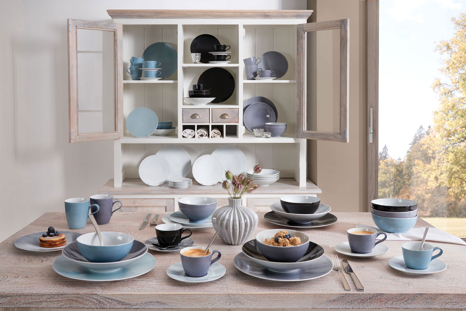 Seltmann Weiden Espressotasse FASHION Verschiedene Geschirrteile in einem offenen Schrank und auf einem Tisch, darunter Tassen, Teller und Schüsseln in verschiedenen Farben und Größen, aus der Perspektive eines gedeckten Tisches mit Blick auf den Schrank.