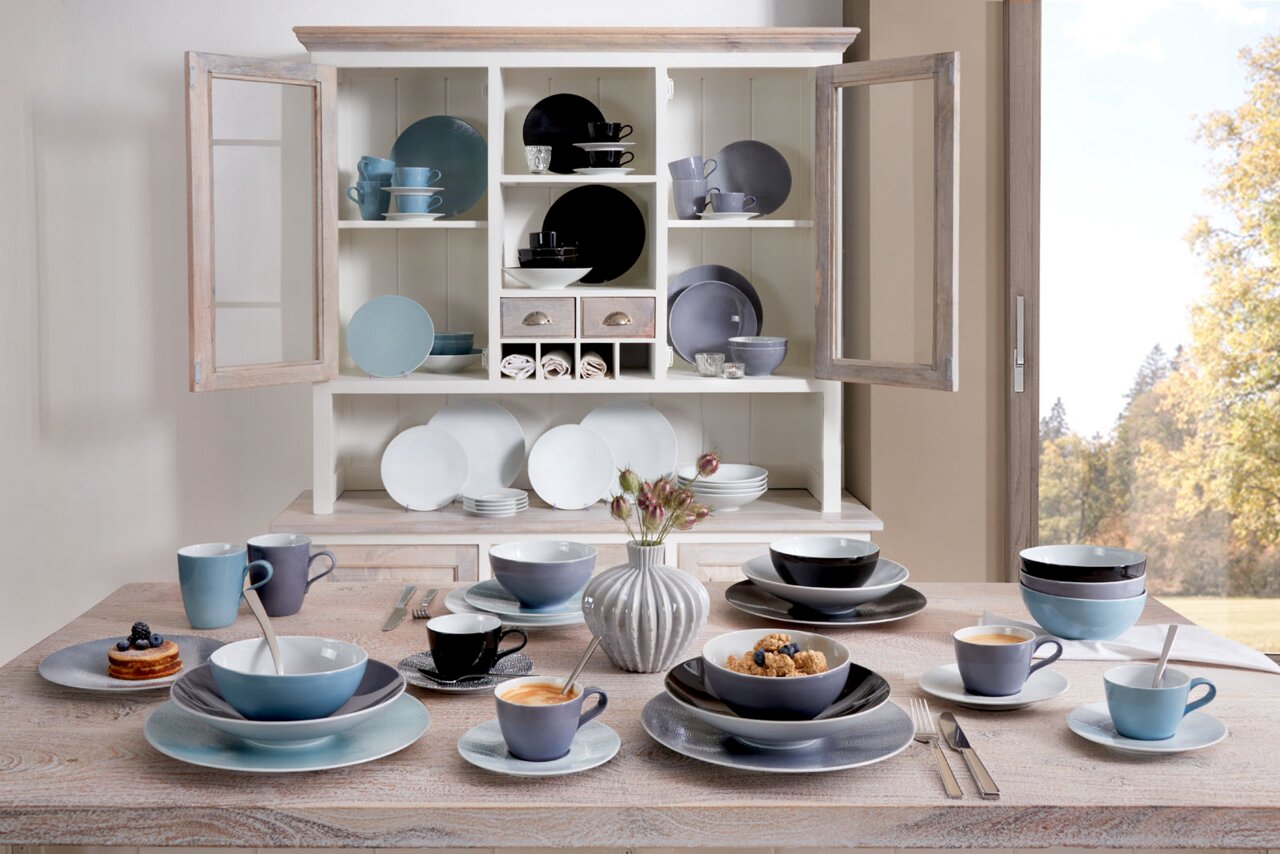 Seltmann Weiden Kaffeetasse Fashion Verschiedene Geschirrteile in einem offenen Schrank und auf einem Tisch, darunter Tassen, Teller und Schüsseln in verschiedenen Farben und Größen, aus der Perspektive eines gedeckten Tisches mit Blick auf den Schrank.