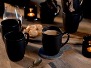 Schwarze Keramik-Kaffeetassen auf Holztisch, eine Tasse mit Kaffee auf Untersetzer, im Hintergrund weitere Becher, Kekse und Kerzenlicht; Perspektive: leicht erhöhte Frontansicht (3/4).
