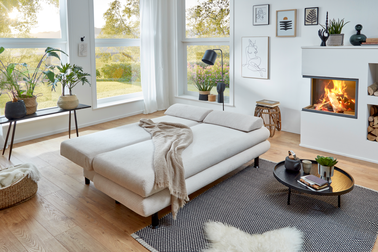 Modernes Funktionssofa Bern in einem gemütlichen Wohnzimmer mit Kamin, aus der Perspektive von vorne links fotografiert.