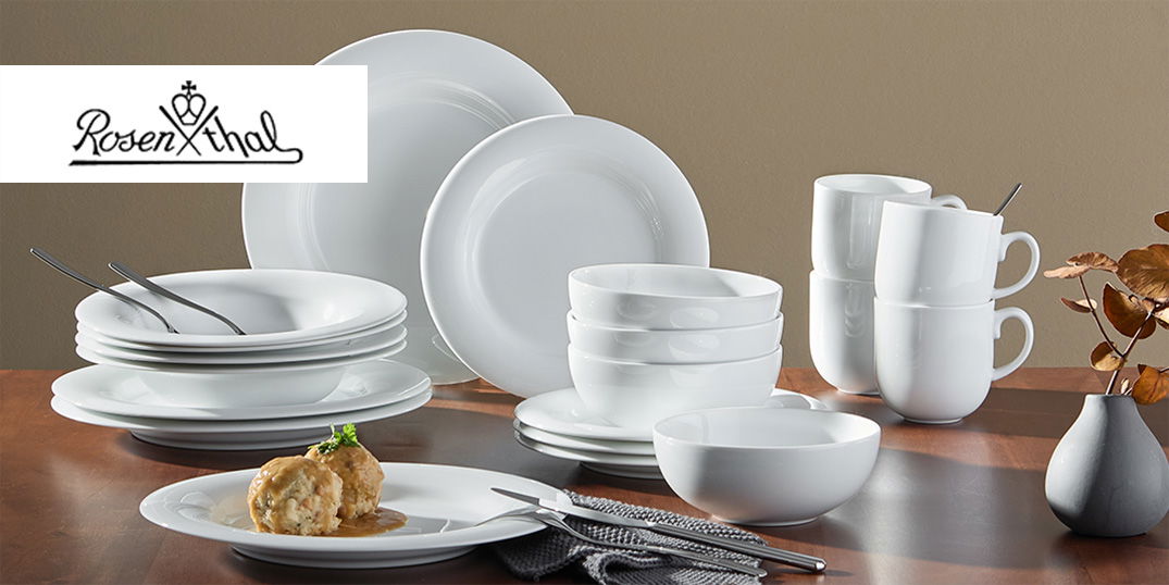 Ein elegantes, weißes Geschirrset mit Tellern, Schüsseln und Tassen steht auf einem gedeckten Holztisch.