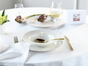 Elegantes Tischgedeck mit weißem Porzellanteller und tiefer Schale, goldfarbenem Besteck und Weinglas mit Weißwein auf weißer Tischdecke, perspektivisch aus leicht erhöhter schräger Vorderansicht
