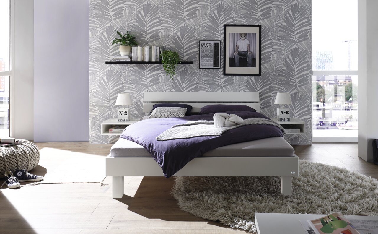 Modernes Schlafzimmer mit einem weißen Holz-Metall-Kopfteil, frontal betrachtet. Das Bett ist mit lila Bettwäsche bezogen und steht auf einem flauschigen Teppich. Im Hintergrund ist eine stilvolle Tapete mit Blattmuster und ein Regal mit Pflanzen und Büch