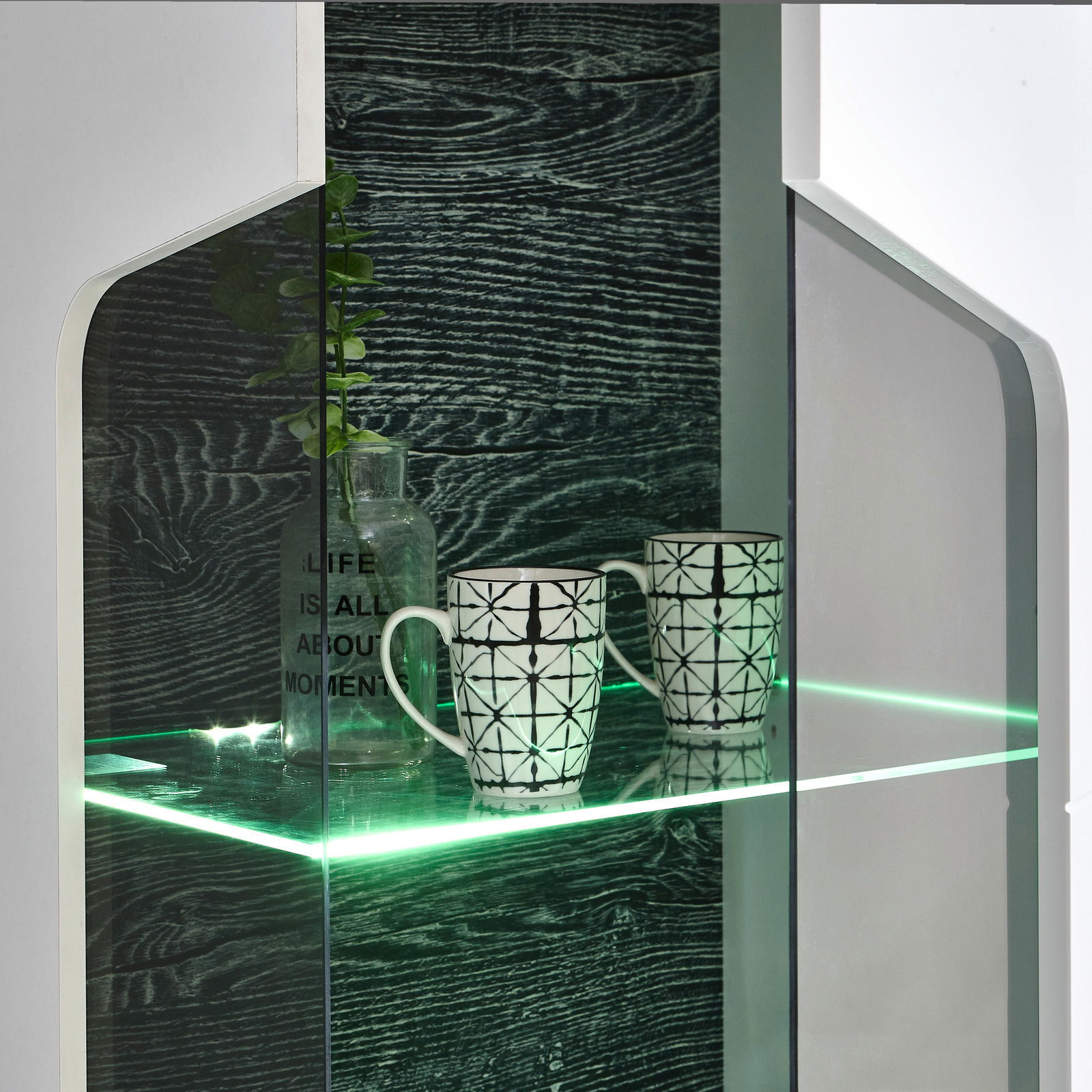 Nahaufnahme einer modernen Vitrine mit Glasregal, beleuchtet mit grünem LED-Licht, zwei gemusterten Tassen und einer dekorativen Flasche. Perspektive von vorne.