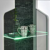 Nahaufnahme einer modernen Vitrine mit Glasregal, beleuchtet mit grünem LED-Licht, zwei gemusterten Tassen und einer dekorativen Flasche. Perspektive von vorne.