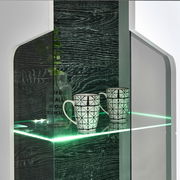 Nahaufnahme einer modernen Vitrine mit Glasregal, beleuchtet mit grünem LED-Licht, zwei gemusterten Tassen und einer dekorativen Flasche. Perspektive von vorne.