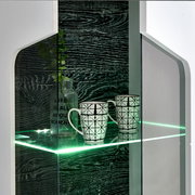 Nahaufnahme einer modernen Vitrine mit Glasregal, beleuchtet mit grünem LED-Licht, zwei gemusterten Tassen und einer dekorativen Flasche. Perspektive von vorne.
