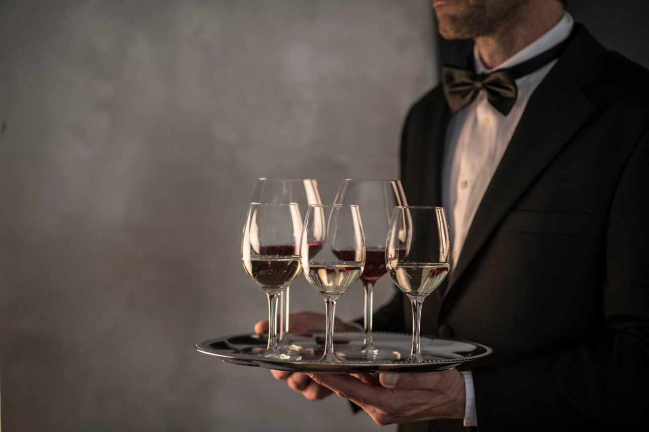SCHOTT ZWIESEL Rotweinglas-Set 4-tlg For You Ein Kellner im Anzug serviert auf einem Tablett vier Rotweingläser, zwei mit Rotwein und zwei mit Weißwein, aus seitlicher Perspektive.