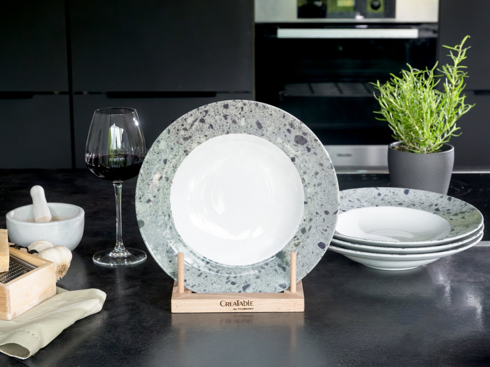 Creatable Pastateller 4-tlg STONE COLLECTION Frontalansicht eines tiefen Porzellantellers in Weiß mit grau gesprenkeltem Rand auf Holzständer, daneben gestapelte Teller in moderner Küche