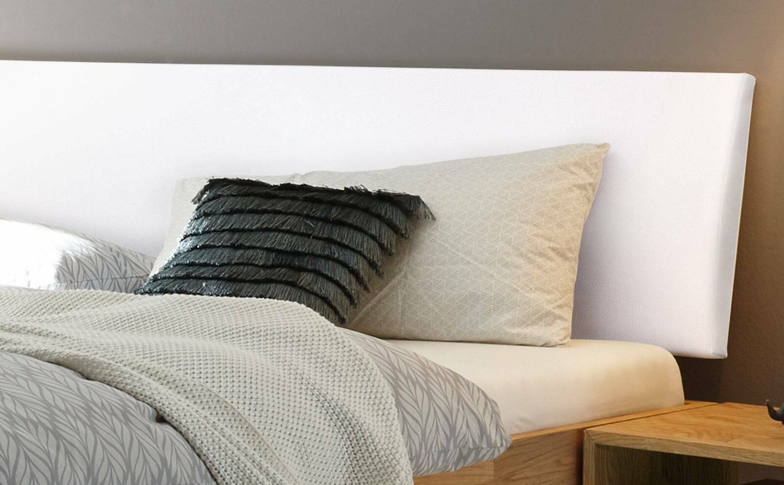 Nahaufnahme eines Bettgestells mit weißem Kopfteil, grauem Kissen mit Fransen und gemustertem Bettbezug, seitliche Perspektive.