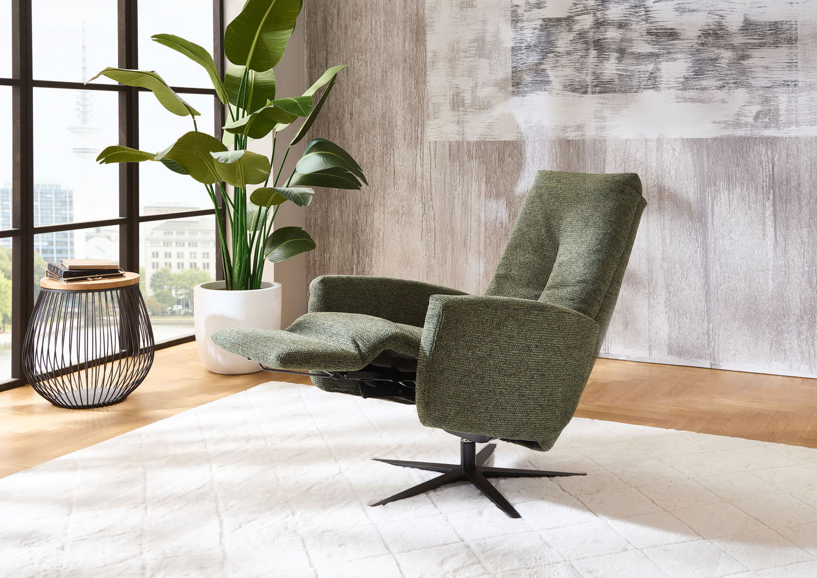 Grüner Relax-Sessel in einem modernen Wohnzimmer, seitliche Perspektive, neben einem großen Fenster und einer Pflanze.