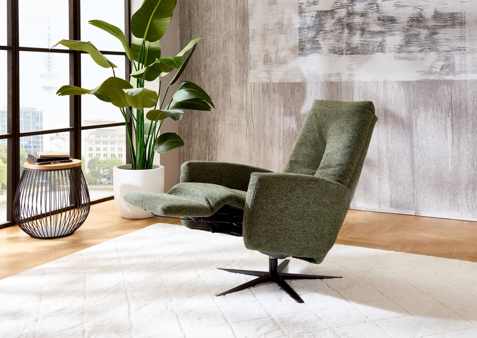 Groene relaxfauteuil in een moderne woonkamer, gezien vanaf de zijkant, naast een groot raam en een plant.