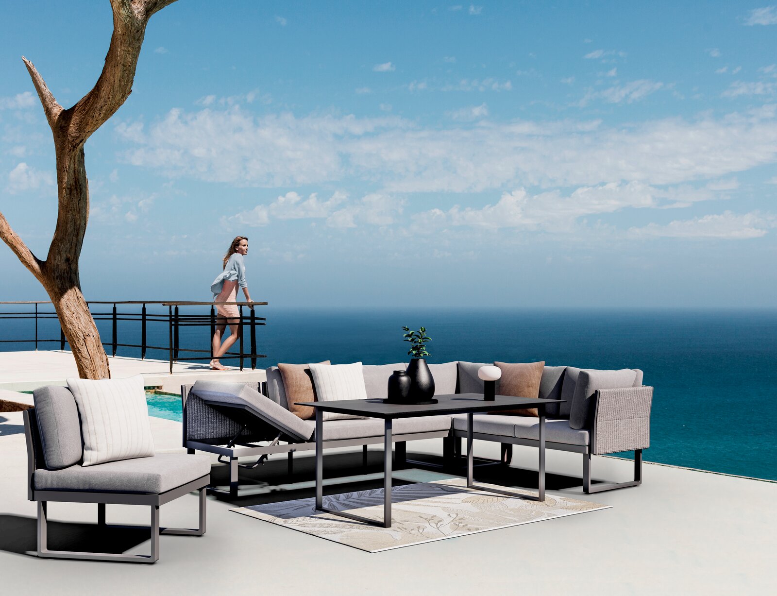 Dieter Knoll Sessel ADELE Moderne Outdoor-Sitzgruppe mit grauen Sofas und Tisch auf einer Terrasse mit Meerblick, seitliche Perspektive.