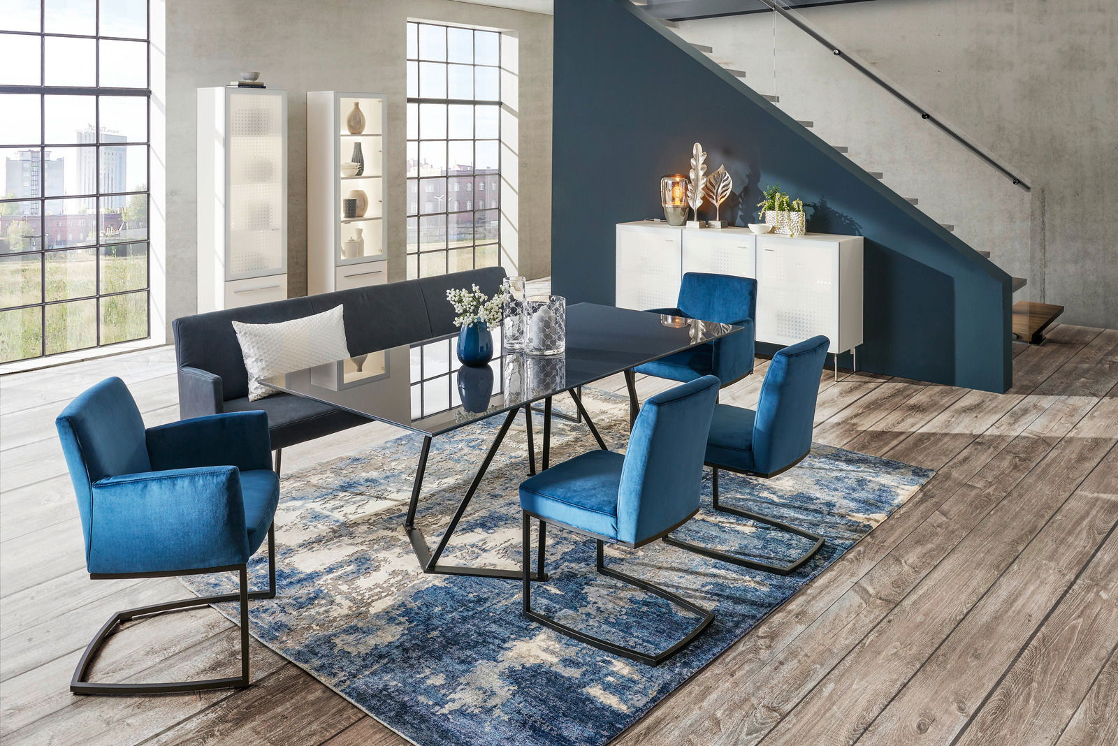 Moderne Esszimmer-Einrichtung mit blauem Freischwinger-Sessel, Esstisch und Sitzbank in einem hellen Raum mit großen Fenstern, aus seitlicher Perspektive.