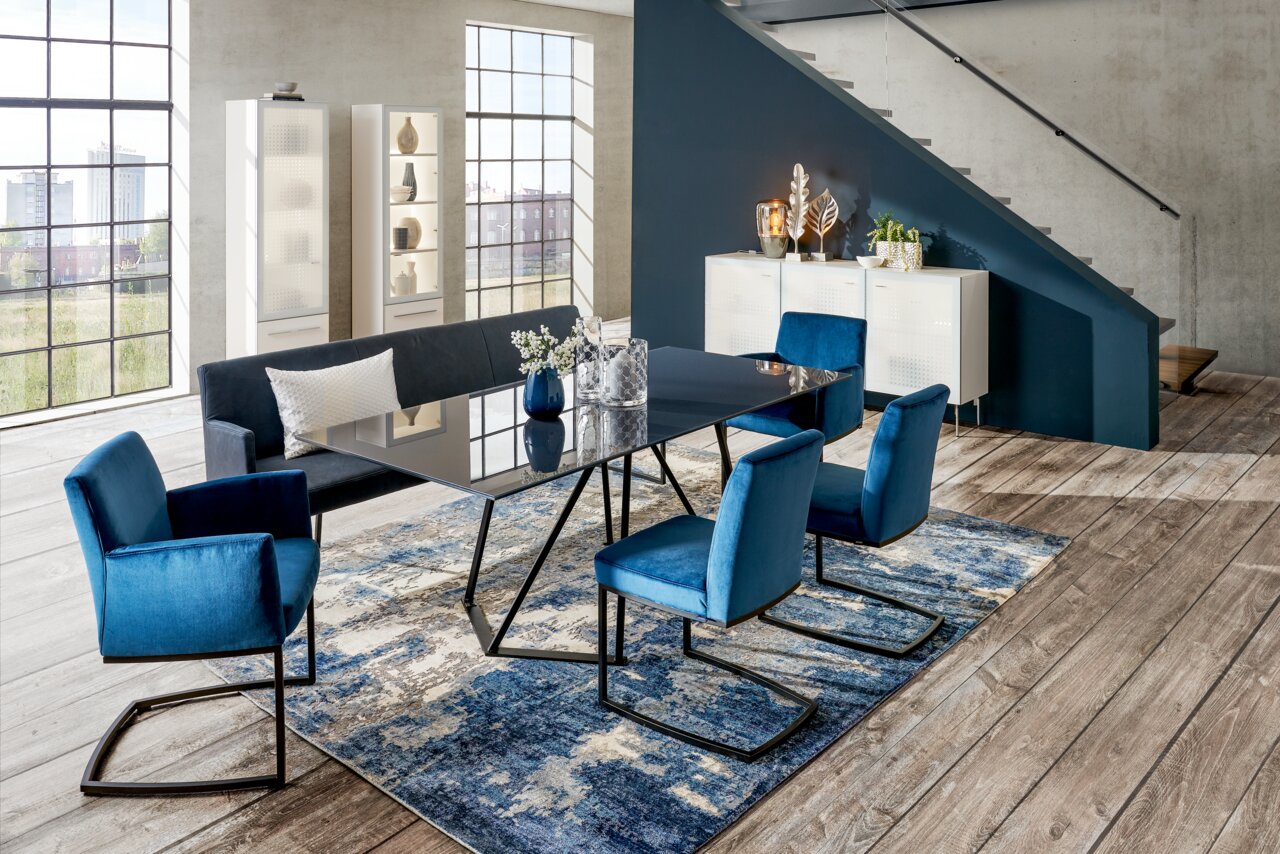 Moderne Esszimmer-Einrichtung mit blauem Freischwinger-Sessel, Esstisch und Sitzbank in einem hellen Raum mit großen Fenstern, aus seitlicher Perspektive.