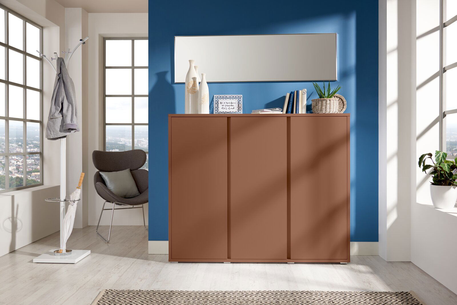 Moderner Schuhschrank in warmem Braunton, frontal fotografiert, vor einer blauen Wand mit Dekorationen und einem Spiegel darüber.
