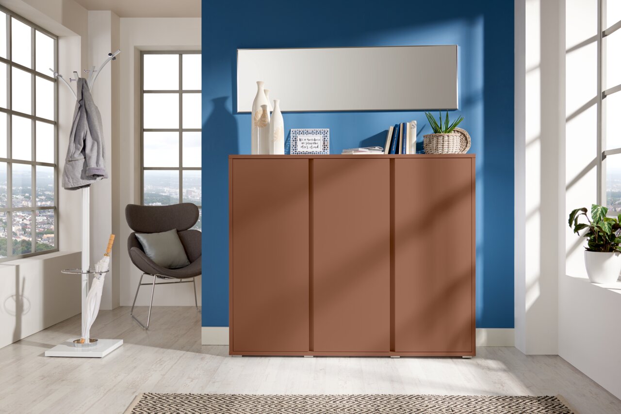 Moderner Schuhschrank in warmem Braunton, frontal fotografiert, vor einer blauen Wand mit Dekorationen und einem Spiegel darüber.