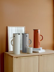 Drei Isolierkannen in verschiedenen Farben, darunter eine in Hellgrau, stehen auf einem Holzschrank vor einer orangefarbenen Wand. Die Kannen sind aus einer seitlichen Perspektive zu sehen.