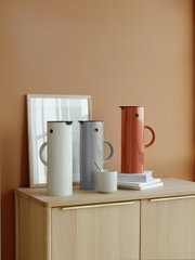 Drie thermoskannen in verschillende kleuren, waaronder een in lichtgrijs, staan op een houten kast voor een oranje muur. De kannen zijn vanuit een zijaanzicht te zien.