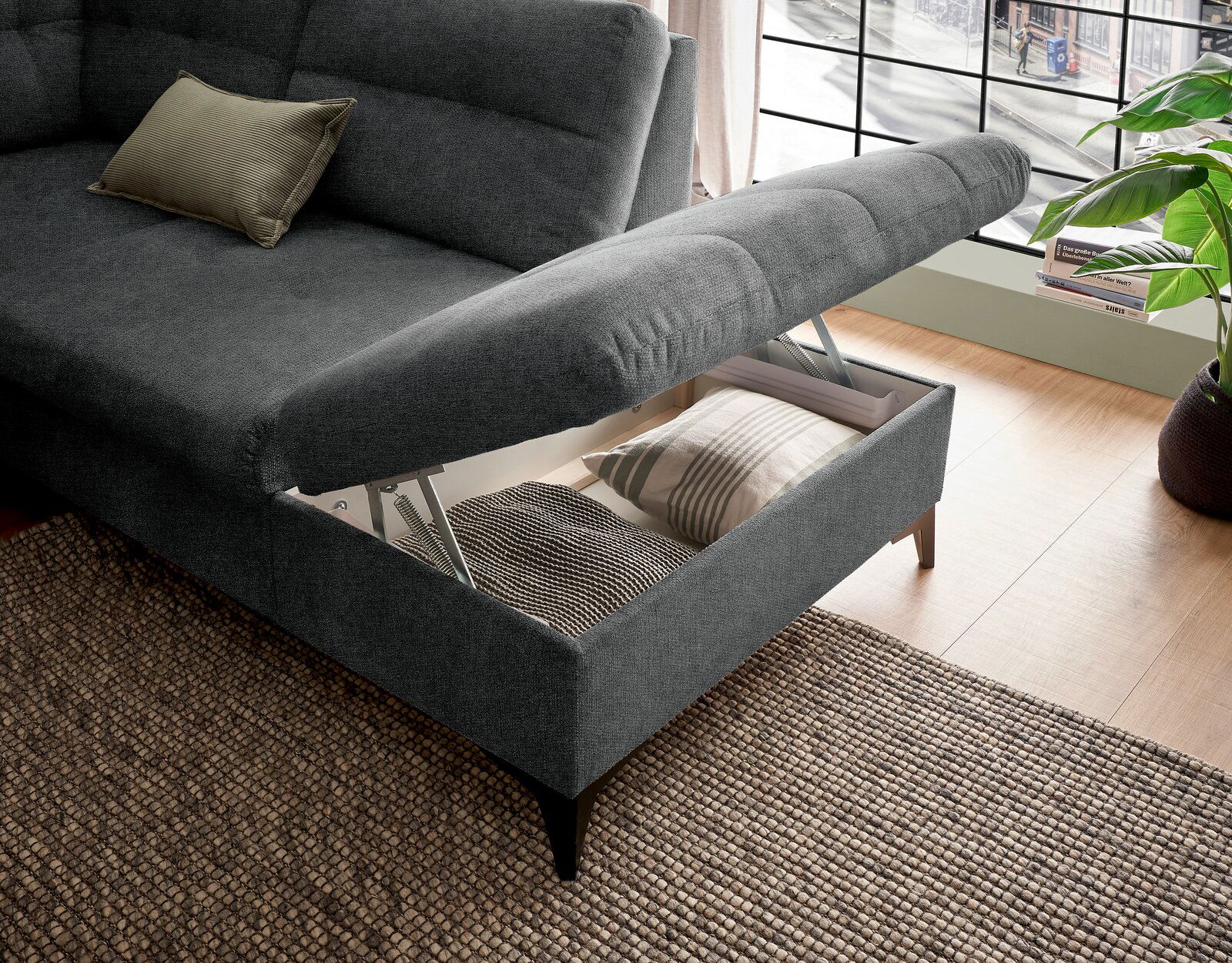 Graues Ecksofa mit hochgeklappter Sitzfläche, die Stauraum mit Kissen zeigt, aus der Vogelperspektive fotografiert.