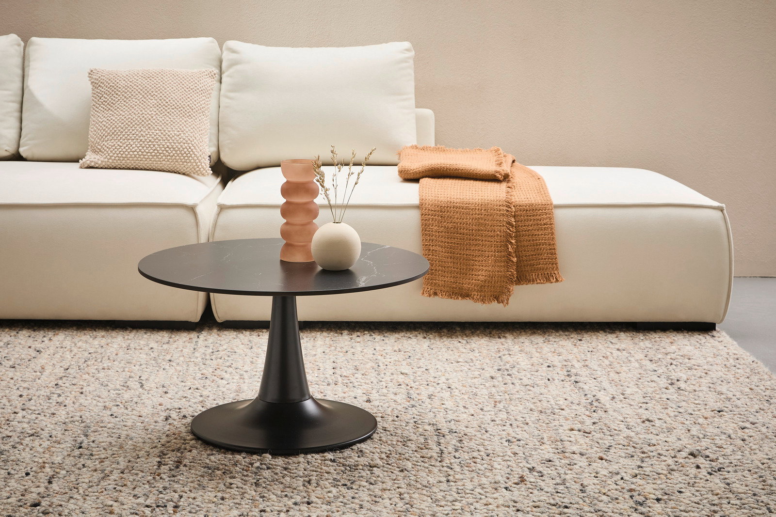 Moderner Couchtisch mit schwarzer runder Tischplatte und konischem Fuß, im Vordergrund eines gemütlichen Wohnzimmers mit beigem Sofa und Teppich, seitliche Perspektive.