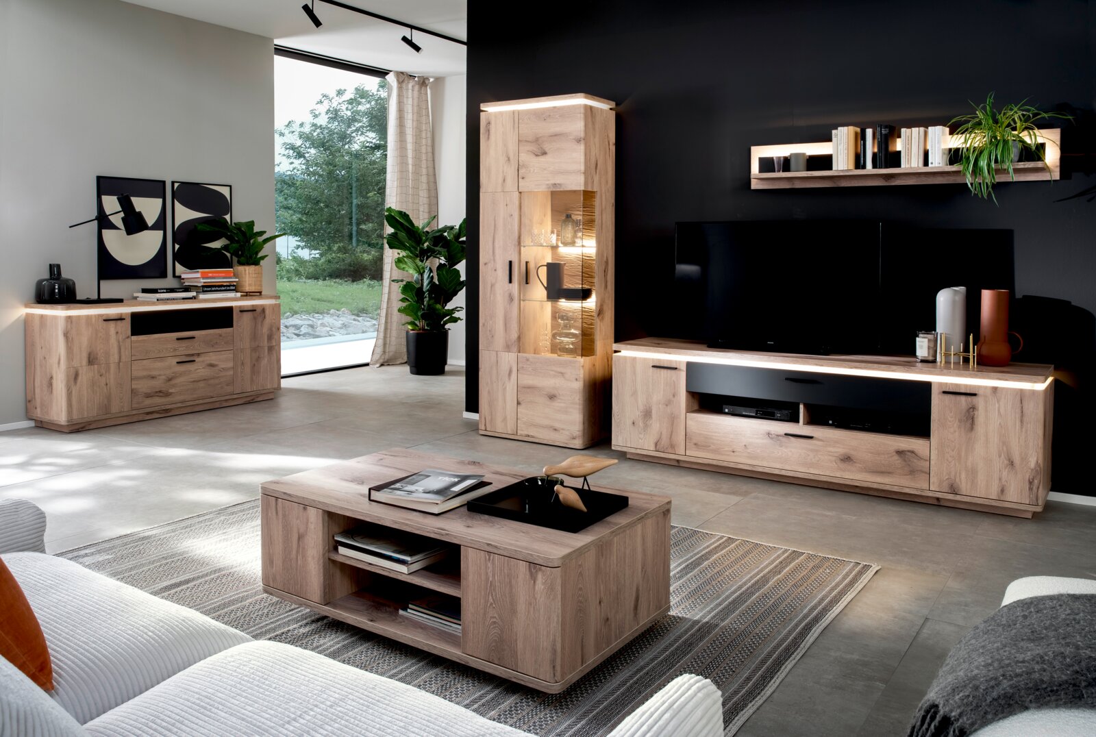 Moderne woonkamer met wandmeubel in eikenlook: tv-lowboard met ledverlichting, vitrinekast, dressoir en salontafel; perspectief: breedbeeldopname van voren, licht van links.