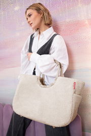 Frau mit einer großen, cremefarbenen Tasche aus weichem Material, die über der Schulter getragen wird. Die Tasche hat runde Griffe und ein rotes Logo. Seitliche Perspektive.