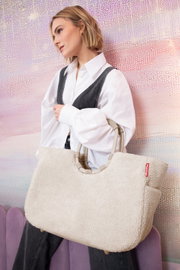 Vrouw met een grote, crèmekleurige tas van zacht materiaal, die over de schouder wordt gedragen. De tas heeft ronde handvatten en een rood logo. Zijaanzicht.