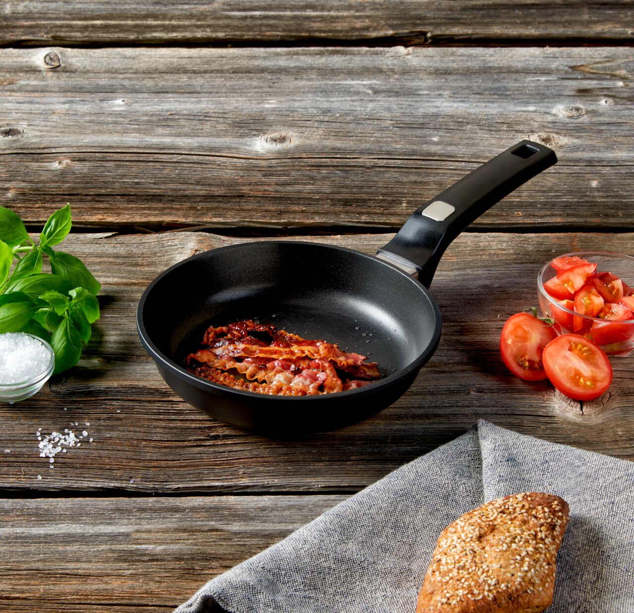 Homeware Pfanne 20 cm Pfanne mit gebratenem Speck auf einem Holztisch, umgeben von frischen Tomaten, Basilikum und einem Brötchen. Perspektive von oben.