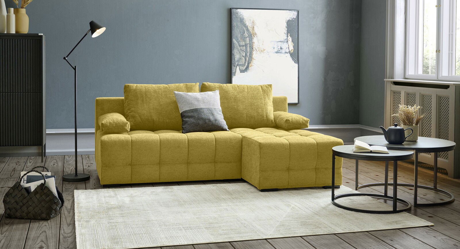 Gelbes Ecksofa MEZZO mit Ottomane rechts, frontal fotografiert in modernem Wohnzimmer mit grauer Wand, Stehlampe und Couchtischen.