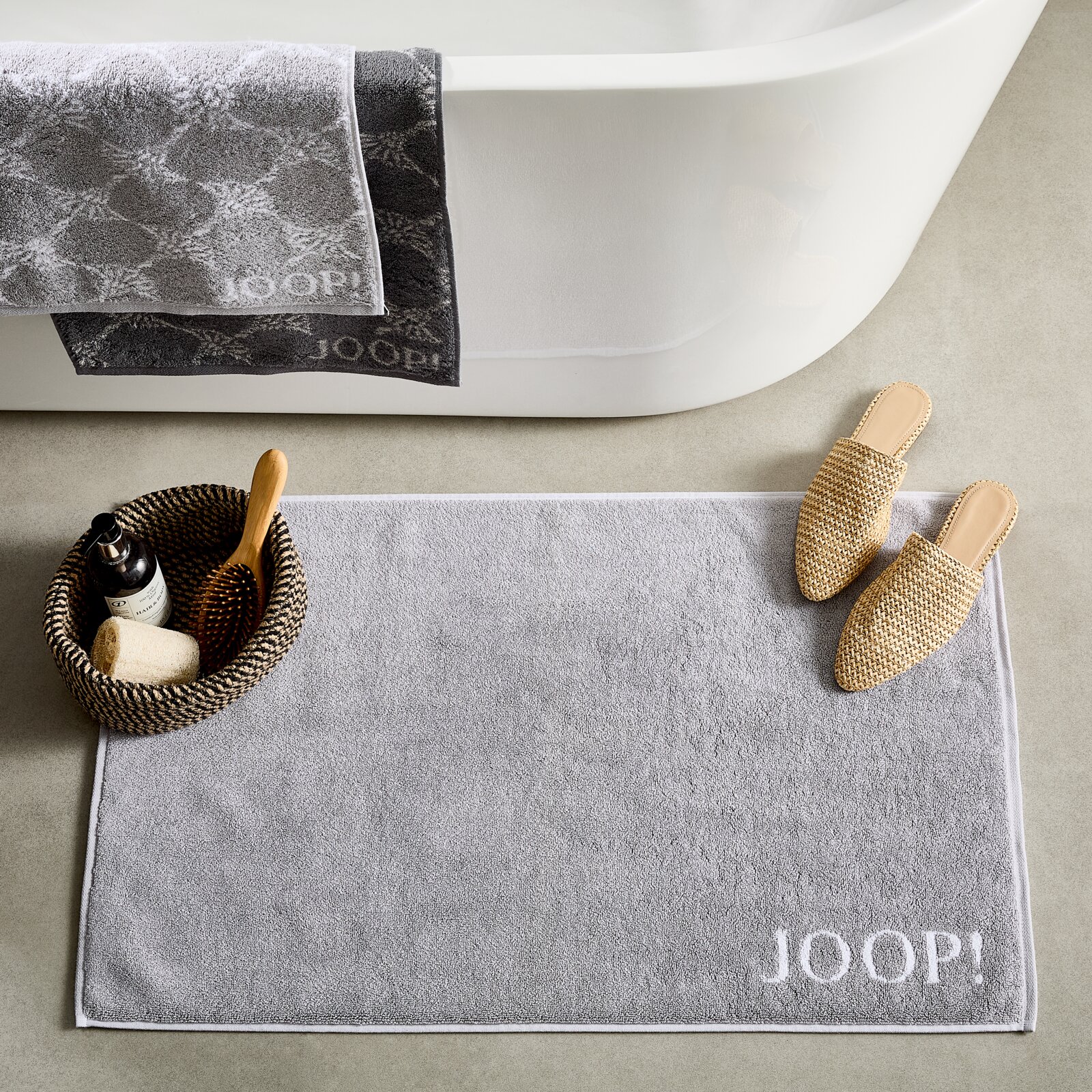Graue Badematte mit dem Schriftzug 'JOOP!' in einer Badezimmerumgebung, von oben fotografiert. Neben der Matte befinden sich Hausschuhe und ein Korb mit Pflegeprodukten.