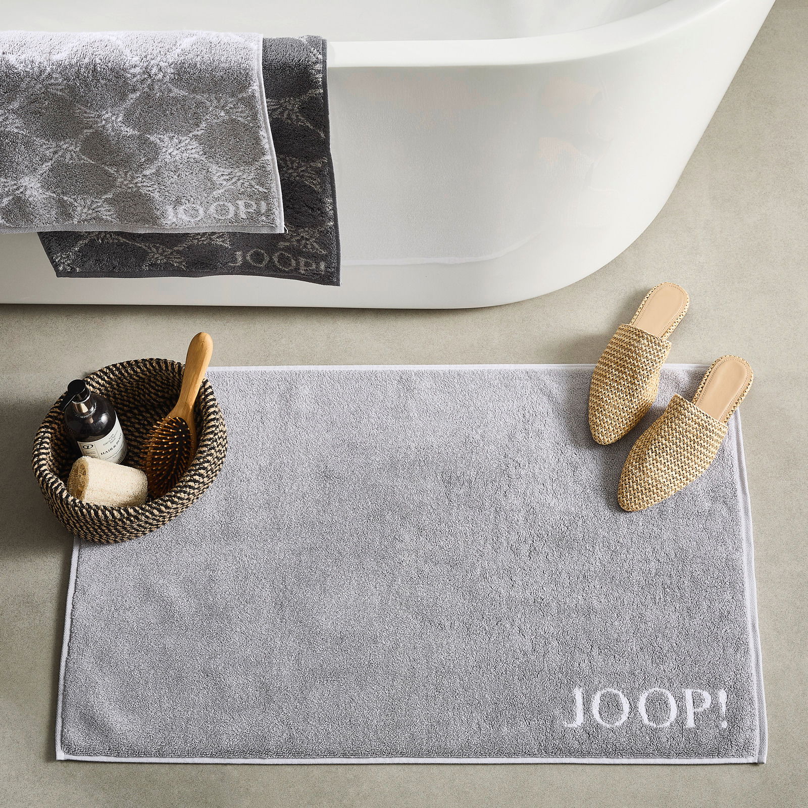 JOOP! Badmat DOUBLEFACE Graue Badematte mit dem Schriftzug 'JOOP!' in einer Badezimmerumgebung, von oben fotografiert. Neben der Matte befinden sich Hausschuhe und ein Korb mit Pflegeprodukten.