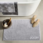 Graue Badematte mit dem Schriftzug 'JOOP!' in einer Badezimmerumgebung, von oben fotografiert. Neben der Matte befinden sich Hausschuhe und ein Korb mit Pflegeprodukten.