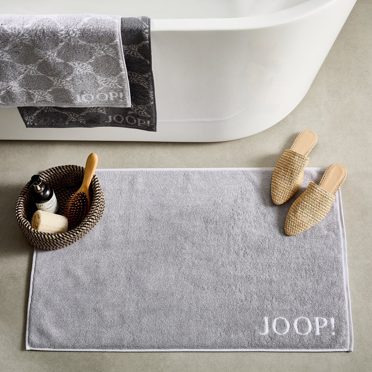 JOOP! Badematte DOUBLEFACE Graue Badematte mit dem Schriftzug 'JOOP!' in einer Badezimmerumgebung, von oben fotografiert. Neben der Matte befinden sich Hausschuhe und ein Korb mit Pflegeprodukten.