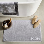 Grijze badmat met het opschrift 'JOOP!' in een badkameromgeving, gefotografeerd van bovenaf. Naast de mat staan pantoffels en een mand met verzorgingsproducten.