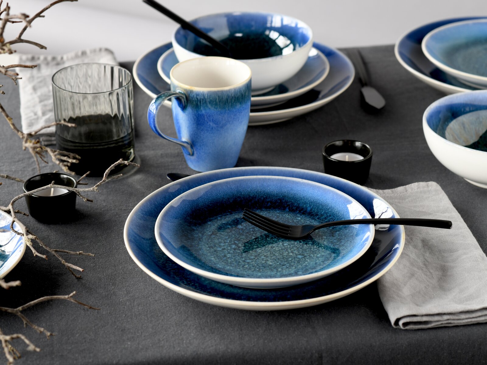 Schlichtes blau-weiß glasiertes Keramikgeschirr-Set mit Teller, Schale und Tasse auf dunkelgrauer Tischdecke, fotografiert aus leicht erhöhter schräger Perspektive (3/4-Ansicht).