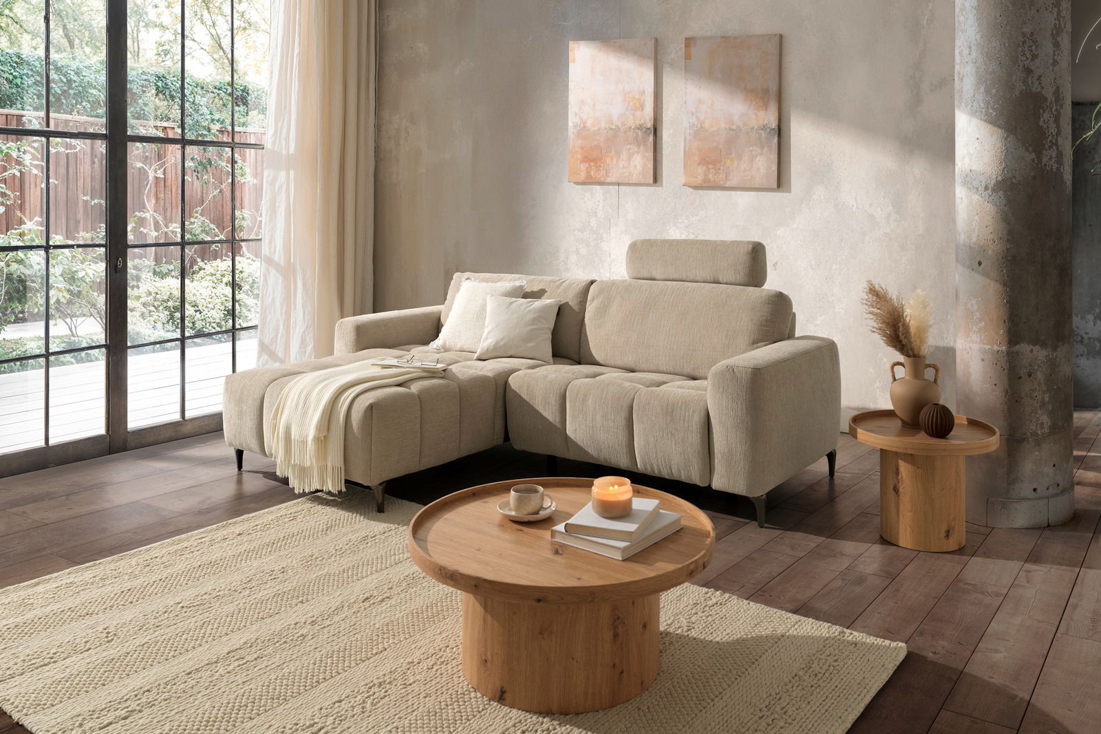 Z2 Ecksofa ROSALY Gemütliche Wohnzimmer-Szene mit einer beigen Rundecke links im Bild, dekoriert mit Kissen und einer Decke, im Hintergrund große Fenster mit Blick ins Grüne.