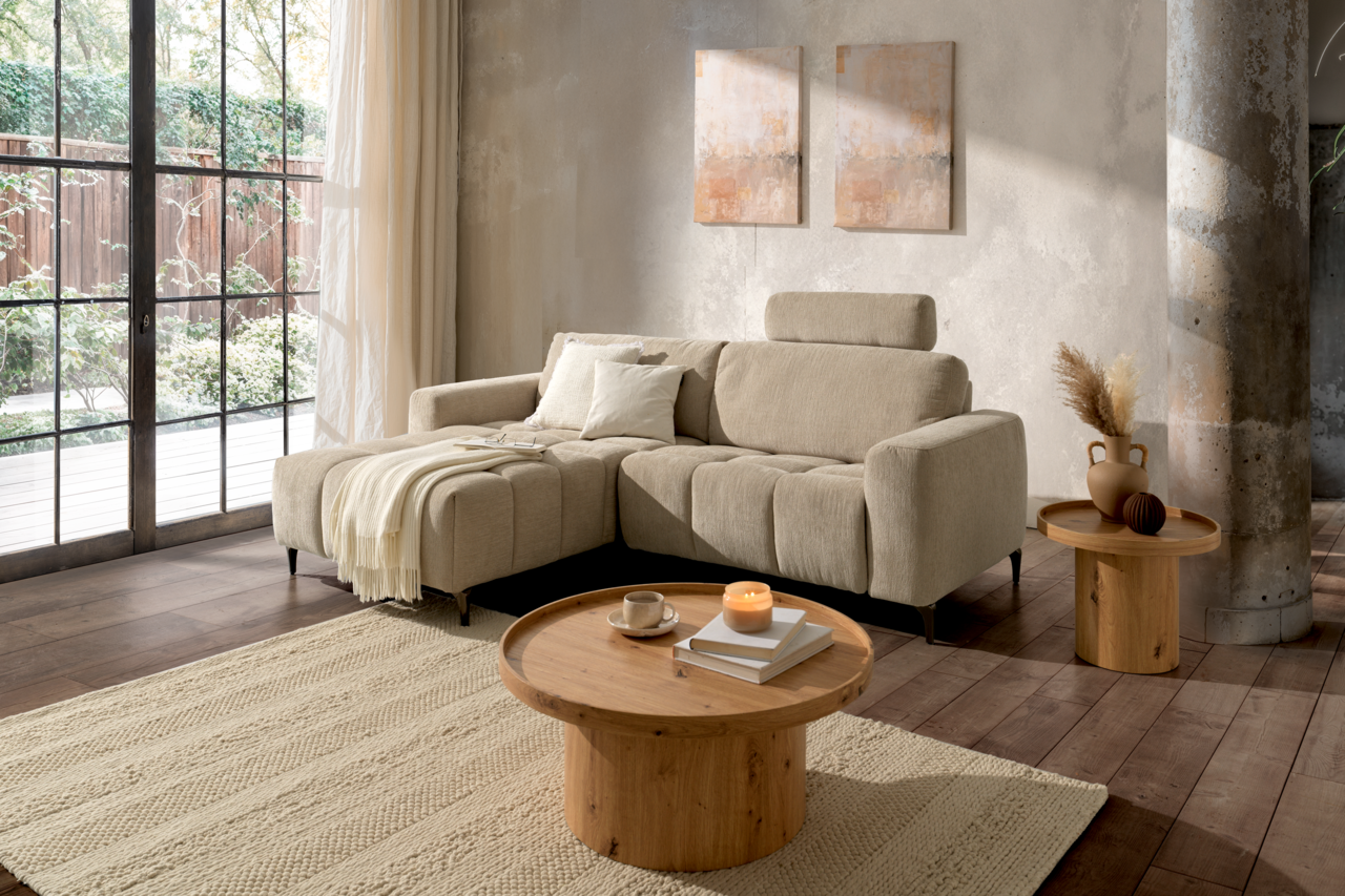 Z2 Ecksofa ROSALY Gemütliche Wohnzimmer-Szene mit einer beigen Rundecke links im Bild, dekoriert mit Kissen und einer Decke, im Hintergrund große Fenster mit Blick ins Grüne.