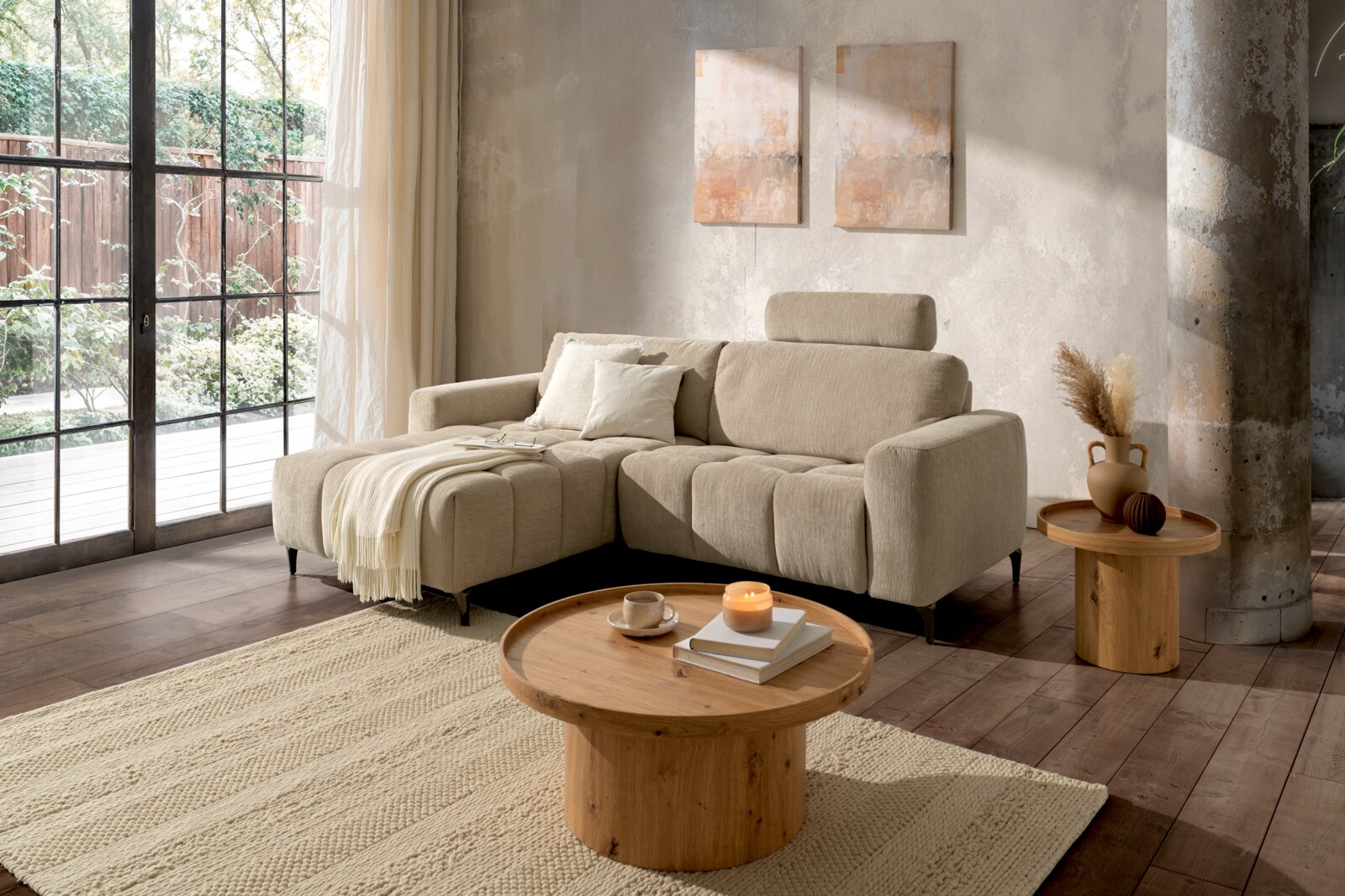 Z2 Ecksofa ROSALY Gemütliche Wohnzimmer-Szene mit einer beigen Rundecke links im Bild, dekoriert mit Kissen und einer Decke, im Hintergrund große Fenster mit Blick ins Grüne.