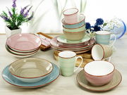 Kombiservice Samba 20-tlg. in verschiedenen Pastellfarben, bestehend aus Tellern, Schalen und Tassen, auf einem Tisch arrangiert, mit Blumen im Hintergrund, aus der Vogelperspektive fotografiert.