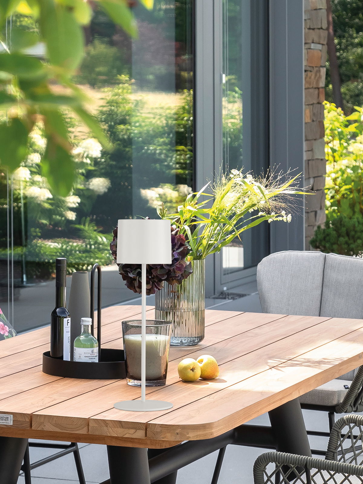 Moderne Akku-Tischleuchte SUN auf einem Holztisch im Freien, umgeben von Pflanzen und Dekorationen, seitliche Perspektive.